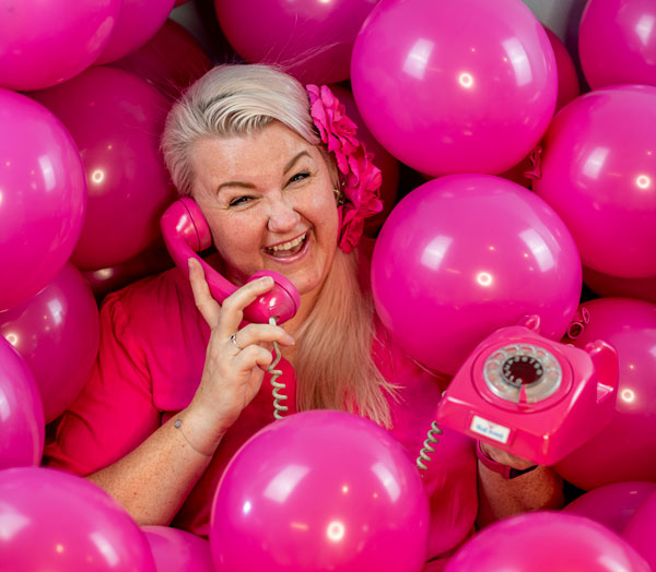 Grunderen og daglig leder i D Lux as, Lill Merete sitter i en haug av rosa sempertex ballonger mens hun holder i en rosa gammeldags telefon med smil om munnen.